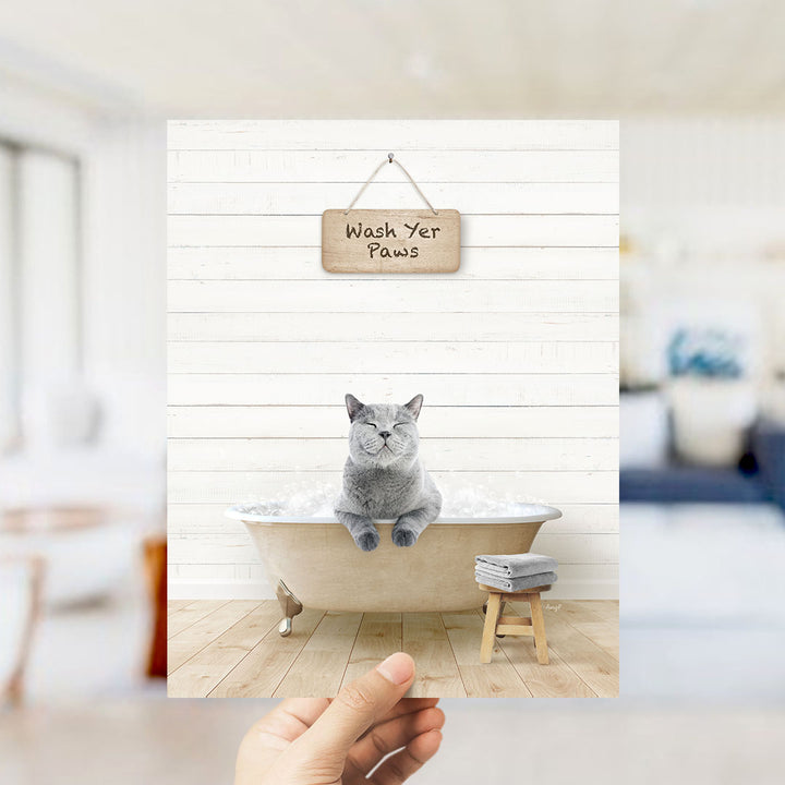 A gray cat sitting in a bathtub filled with bubbles, with a wooden sign above that says "Wash Yer Paws".