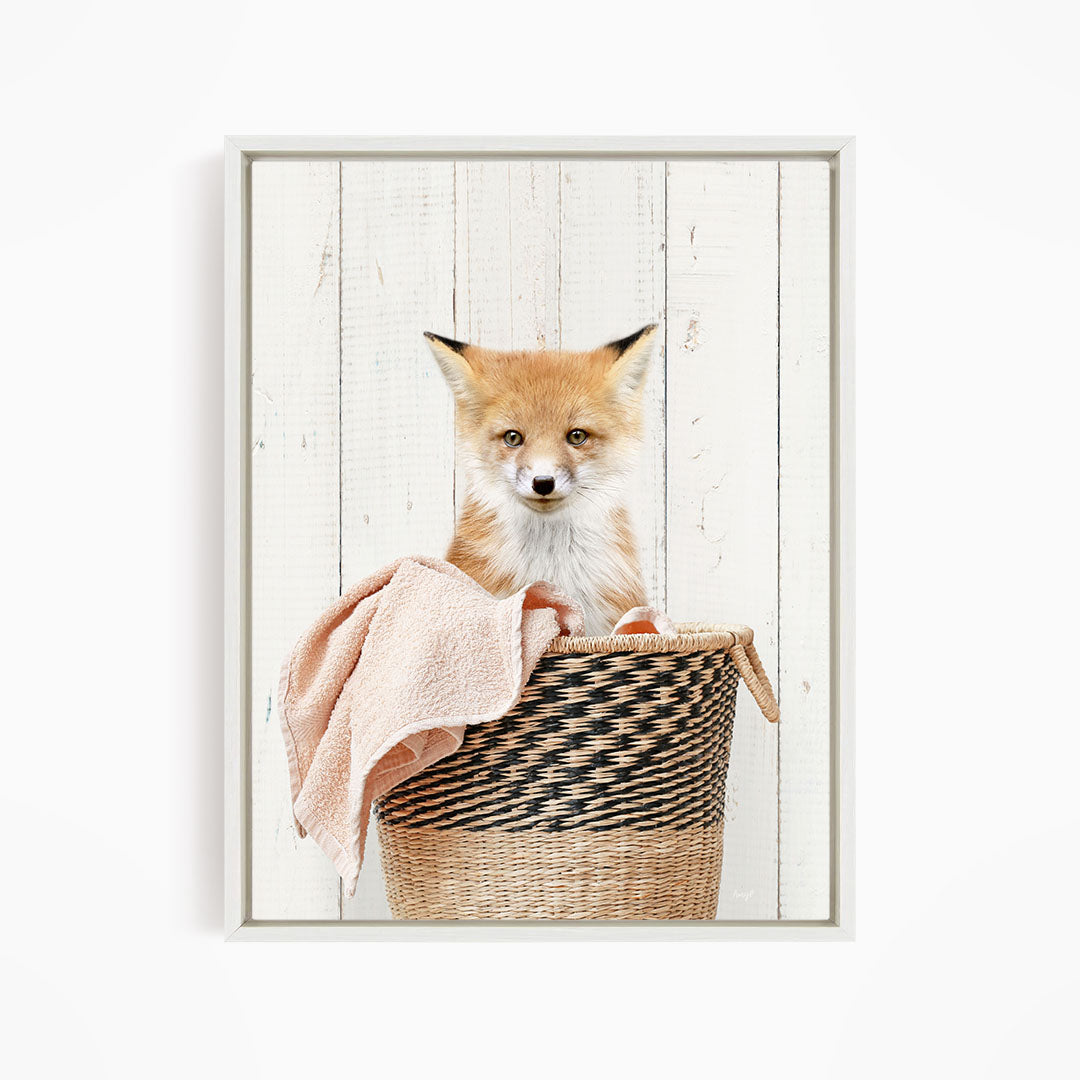 A young fox sits in a woven basket against a white wooden background.