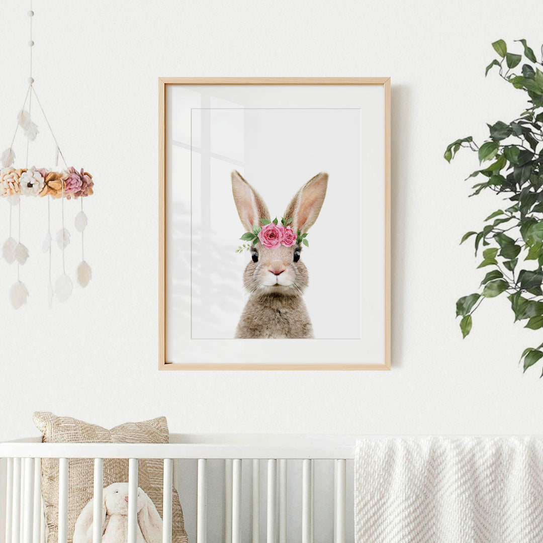 A framed picture of a bunny with a flower crown hangs on a white wall, accompanied by a mobile with flowers and a white crib with a stuffed animal in the corner.