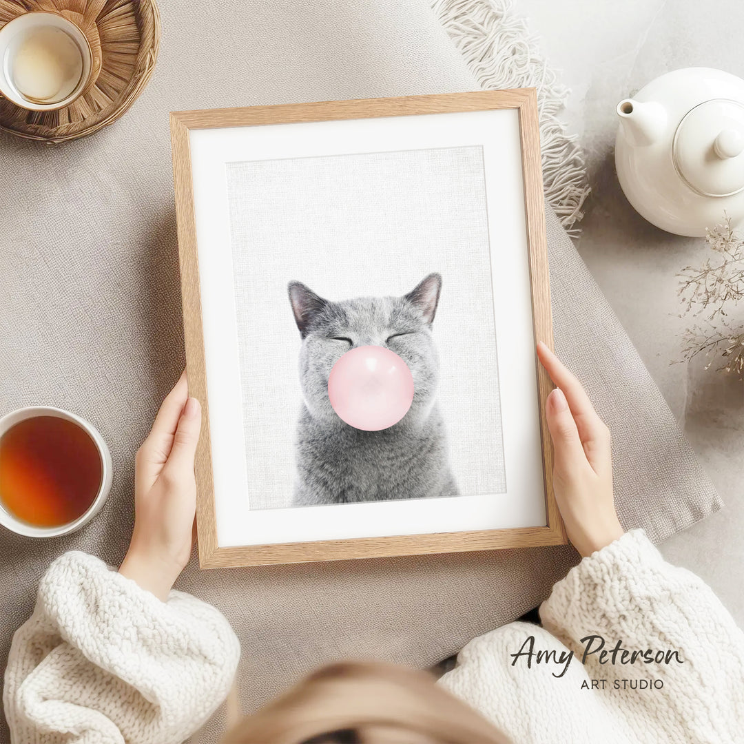 A person is holding a framed picture of a gray cat blowing a bubble with a pink bubble gum bubble.