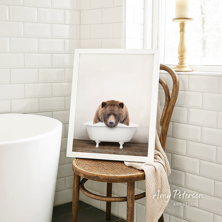 A framed image of a brown bear sitting in a white bathtub, with a towel draped over the back of a wooden chair in a bathroom setting.