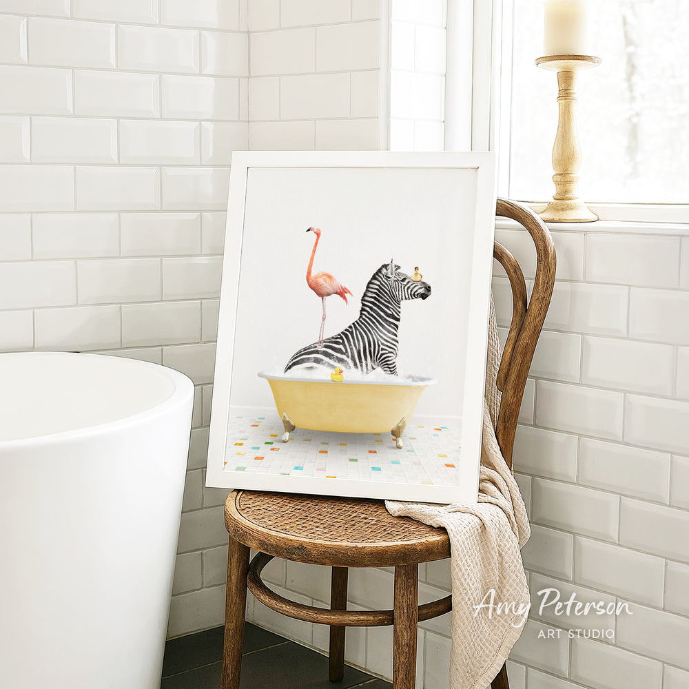 A framed image of a zebra sitting in a bathtub, with a pink flamingo standing on its back, is displayed on a wooden chair in a bathroom setting.