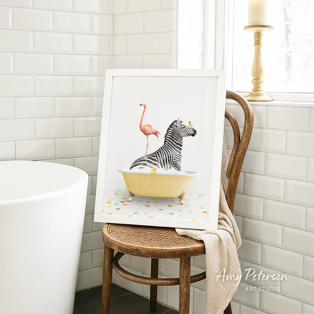 A framed image of a zebra sitting in a bathtub, with a pink flamingo standing on its back, is displayed on a wooden chair in a bathroom setting.