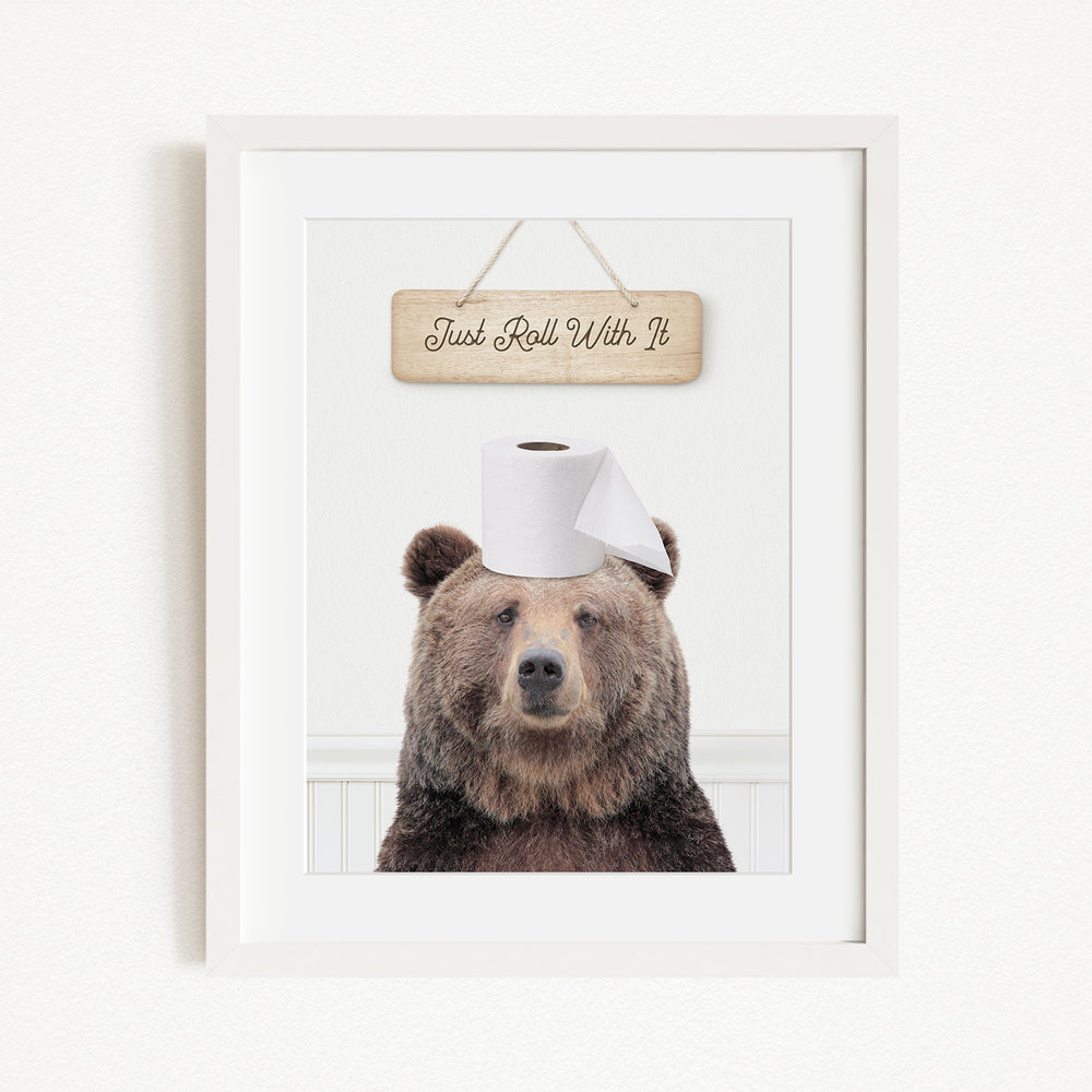 A framed image of a brown bear wearing a toilet paper hat, with a wooden sign above it that says "Just Roll With It".