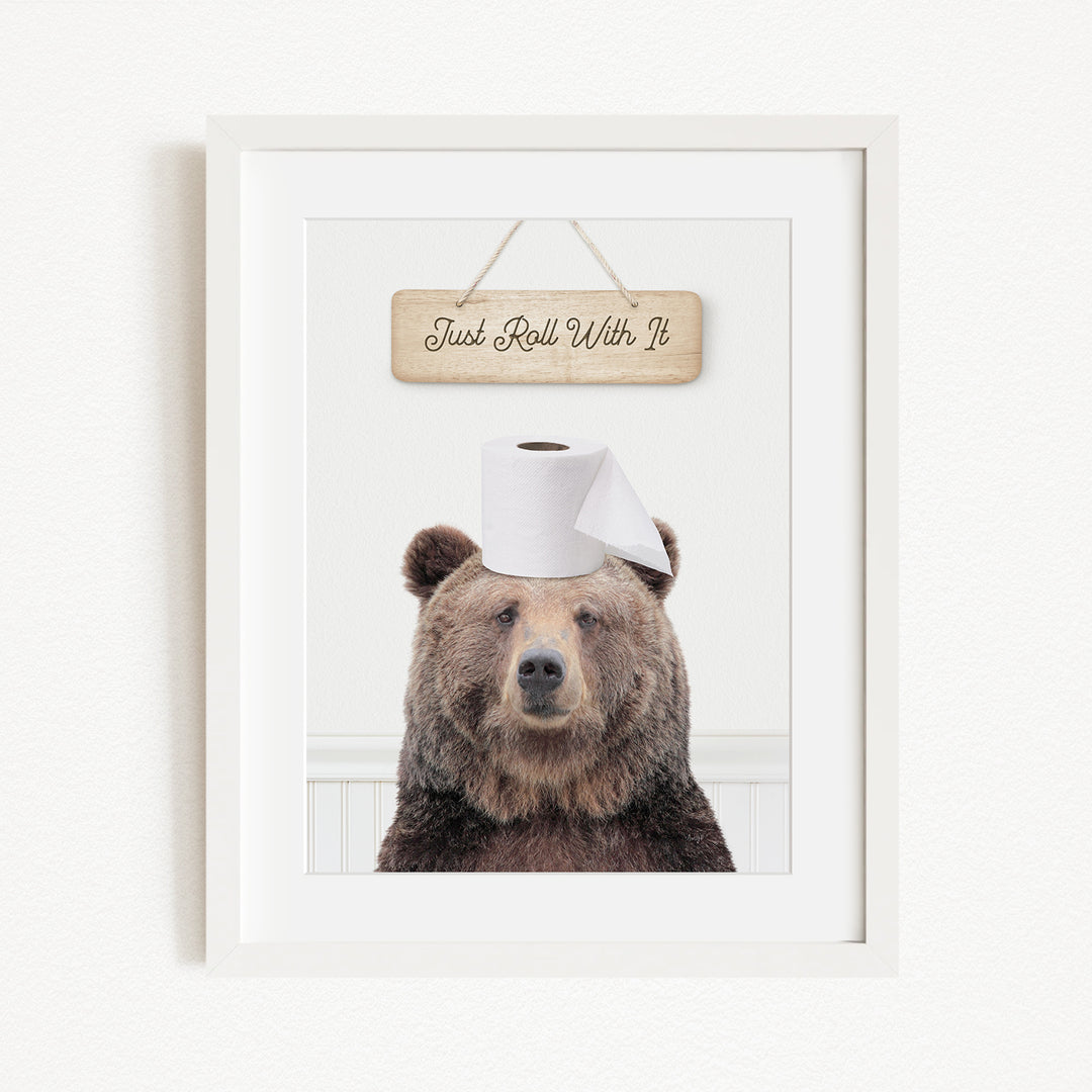 A framed image of a brown bear wearing a toilet paper hat, with a wooden sign above it that says "Just Roll With It".