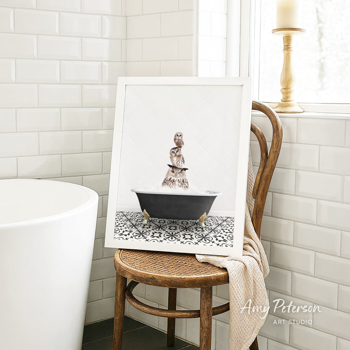 A framed image of a cat sitting in a bathtub, surrounded by a black and white patterned tile floor, is displayed on a wooden chair in a bathroom setting.