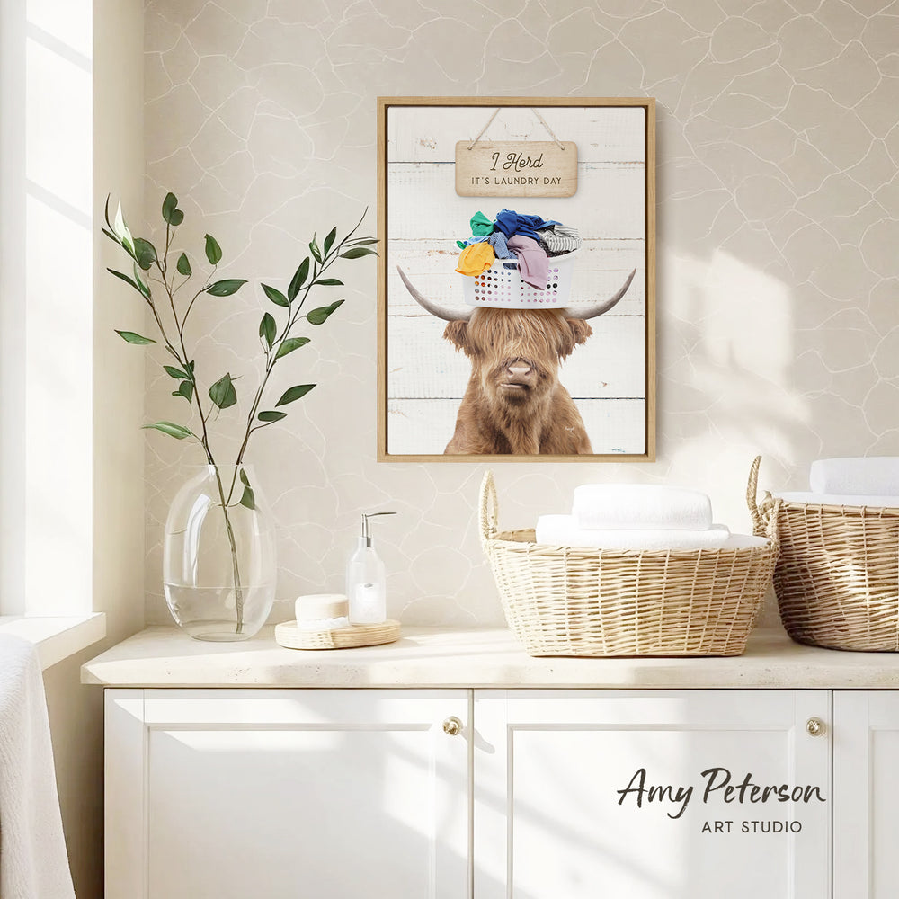 a framed picture of a Highland cow wearing a laundry basket hat, placed on a white cabinet in a bathroom setting.