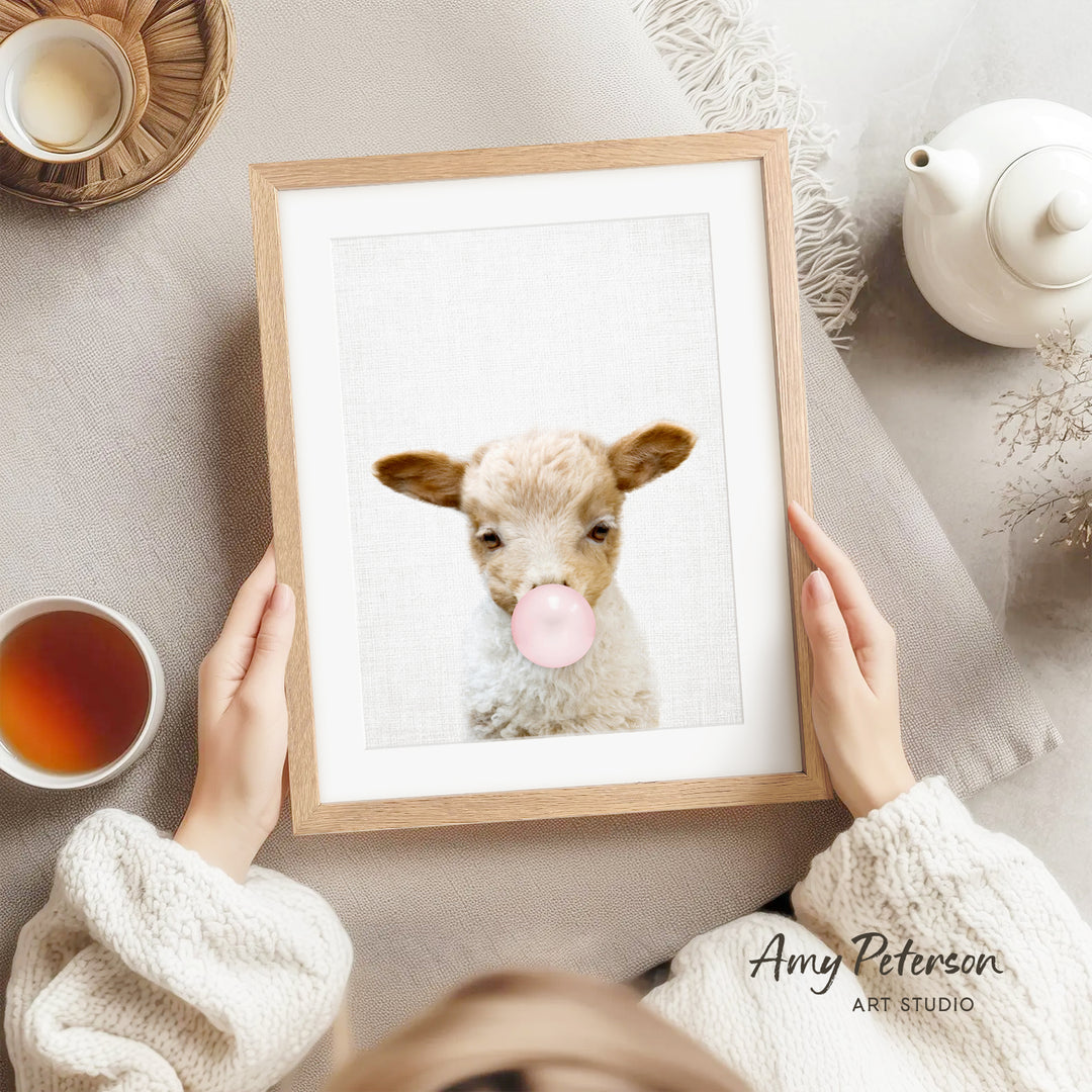 A person is holding a framed picture of a lamb with a pink bubble gum bubble in its mouth, surrounded by various objects such as a teapot, a cup, and a bowl.