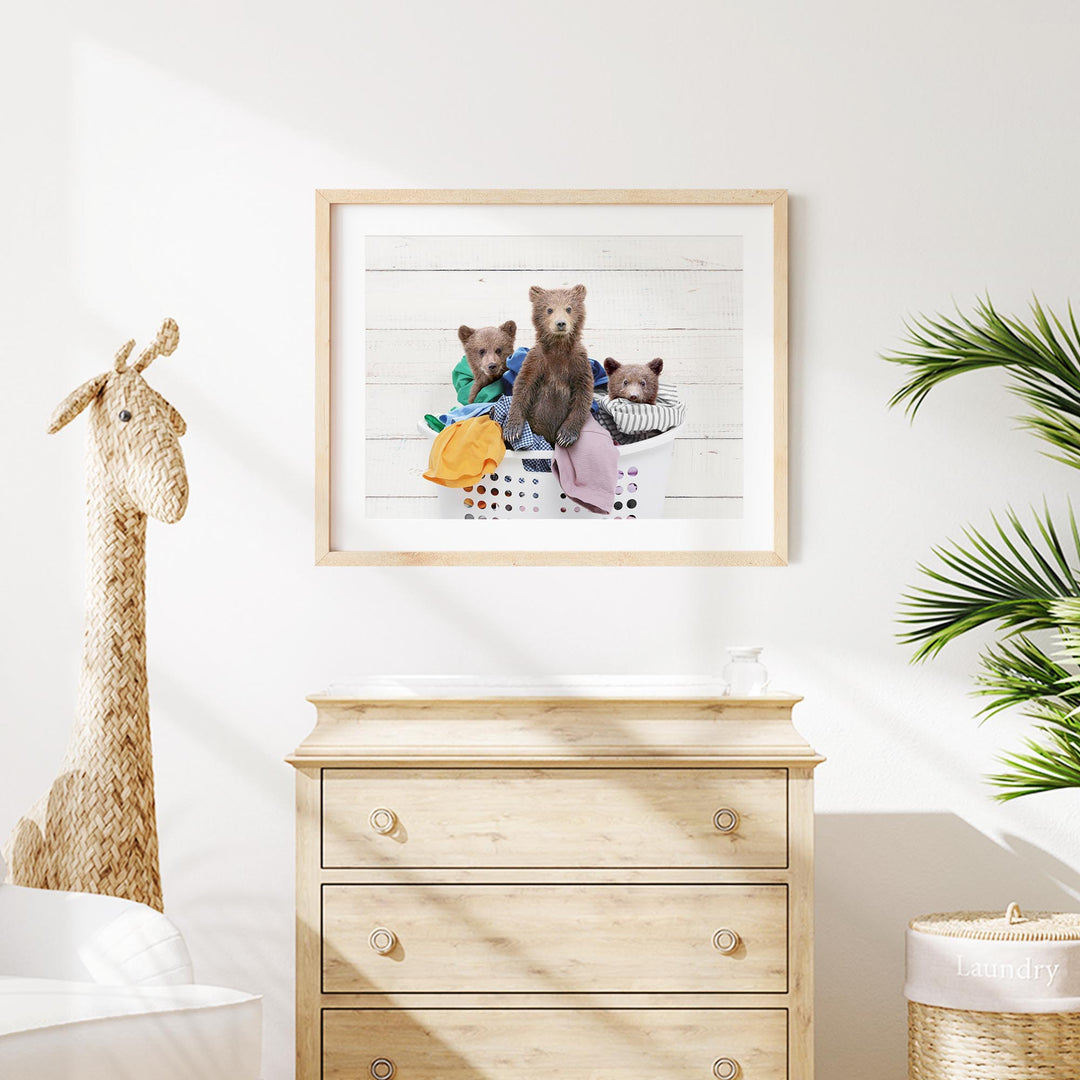 A framed photo of three bears sitting in a laundry basket is displayed on a white wall above a wooden dresser.