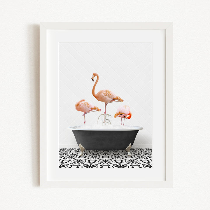 A framed image depicting three pink flamingos standing in a black bathtub filled with water, set against a white background with a black and white patterned floor.