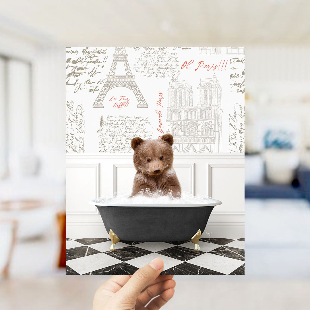 A hand is holding up a framed image of a small brown bear sitting in a bathtub filled with bubbles, with a Paris-themed background featuring the Eiffel Tower and other Parisian landmarks.