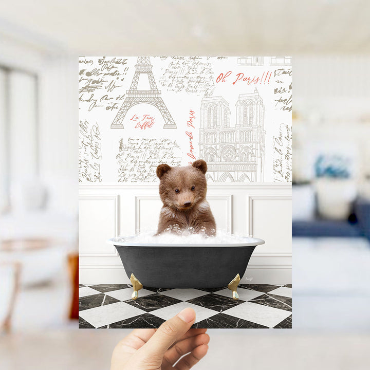 A hand is holding up a framed image of a small brown bear sitting in a bathtub filled with bubbles, with a Paris-themed background featuring the Eiffel Tower and other Parisian landmarks.