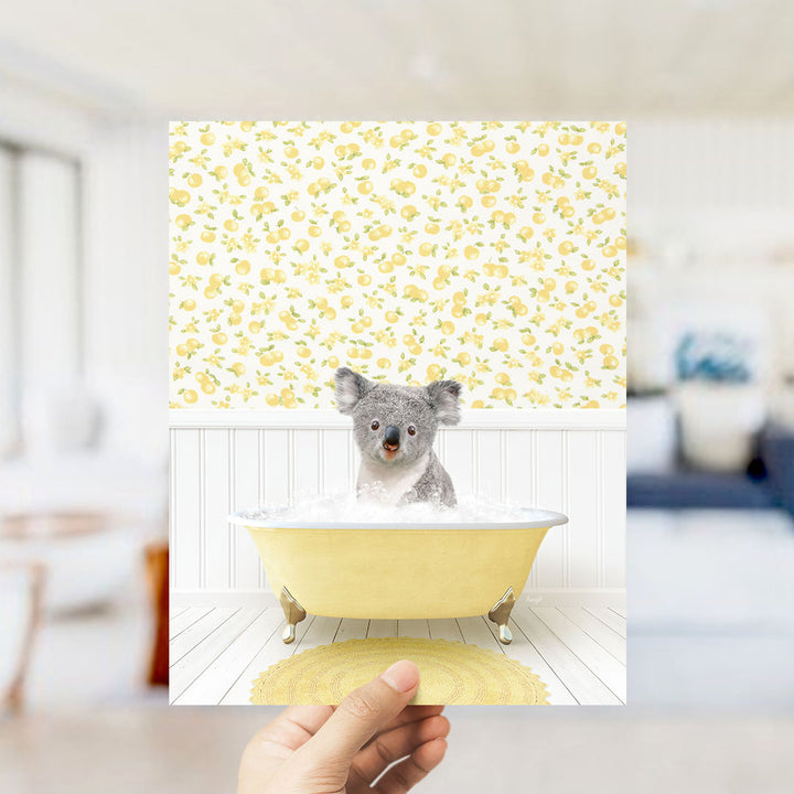 A hand holds up a framed image of a koala bear sitting in a yellow bathtub filled with bubbles, against a yellow and white floral wallpaper.