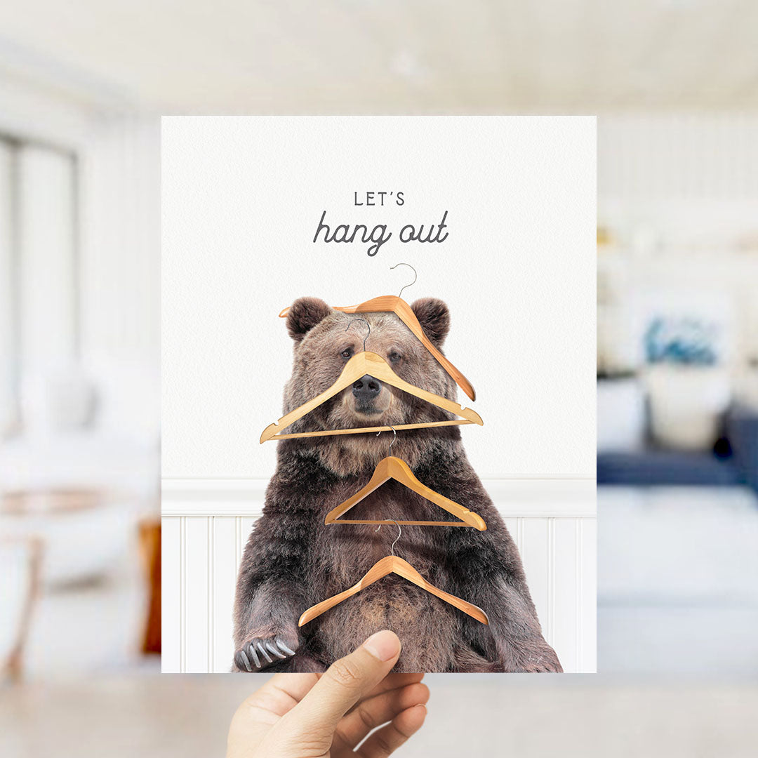 A hand is holding up a card with a bear sitting on a shelf and hanging clothes on it.