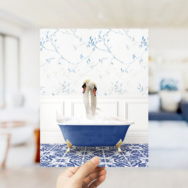 A hand holds up a framed image of two swans sitting in a blue bathtub, with a floral wallpaper in the background.