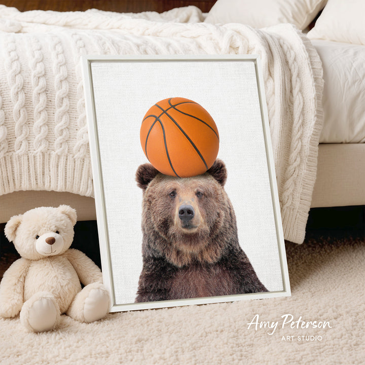 A framed image of a brown bear with a basketball on its head, placed on a bed next to a stuffed teddy bear.