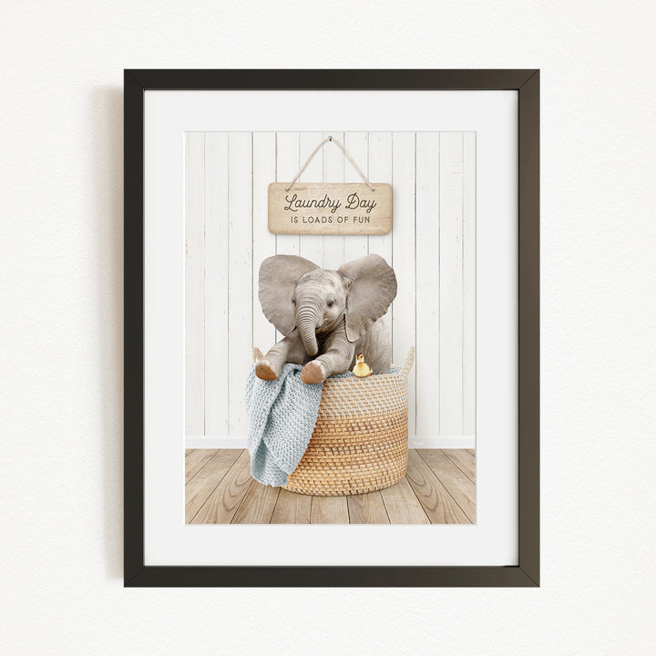 A framed image of a baby elephant sitting in a woven basket, with a sign above that says "Laundry Day is loads of fun".