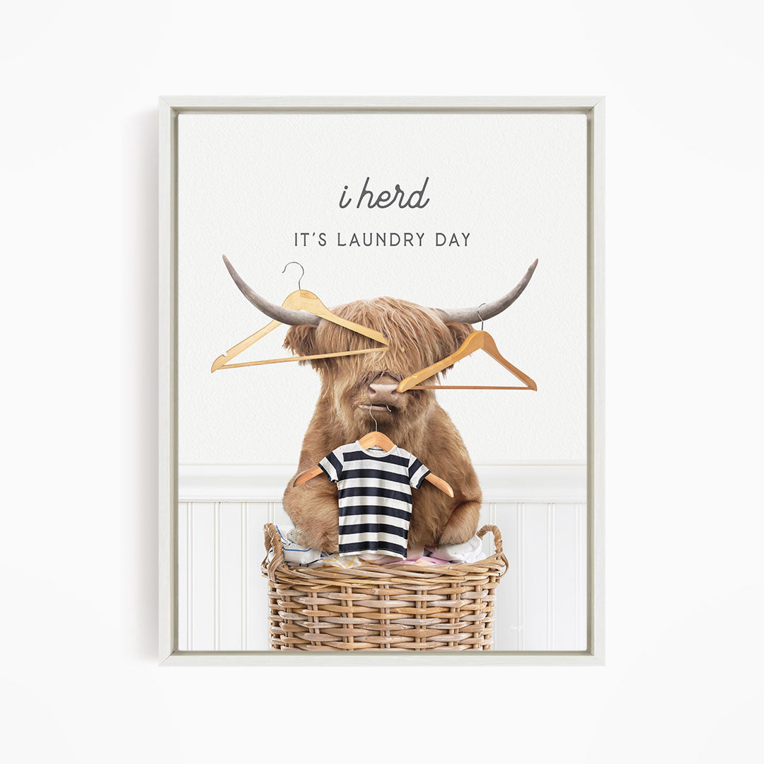A brown cow with horns sits in a wicker basket, holding a pair of clothes hangers. The text above the cow reads "i herd IT'S Laundry Day".