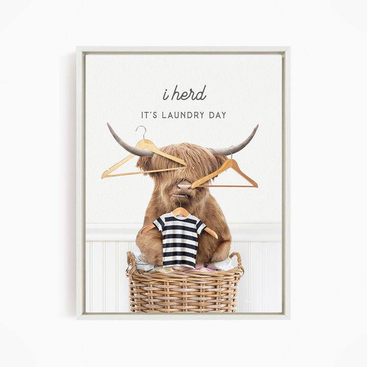 A brown cow with horns sits in a wicker basket, holding a pair of clothes hangers. The text above the cow reads "i herd IT'S Laundry Day".