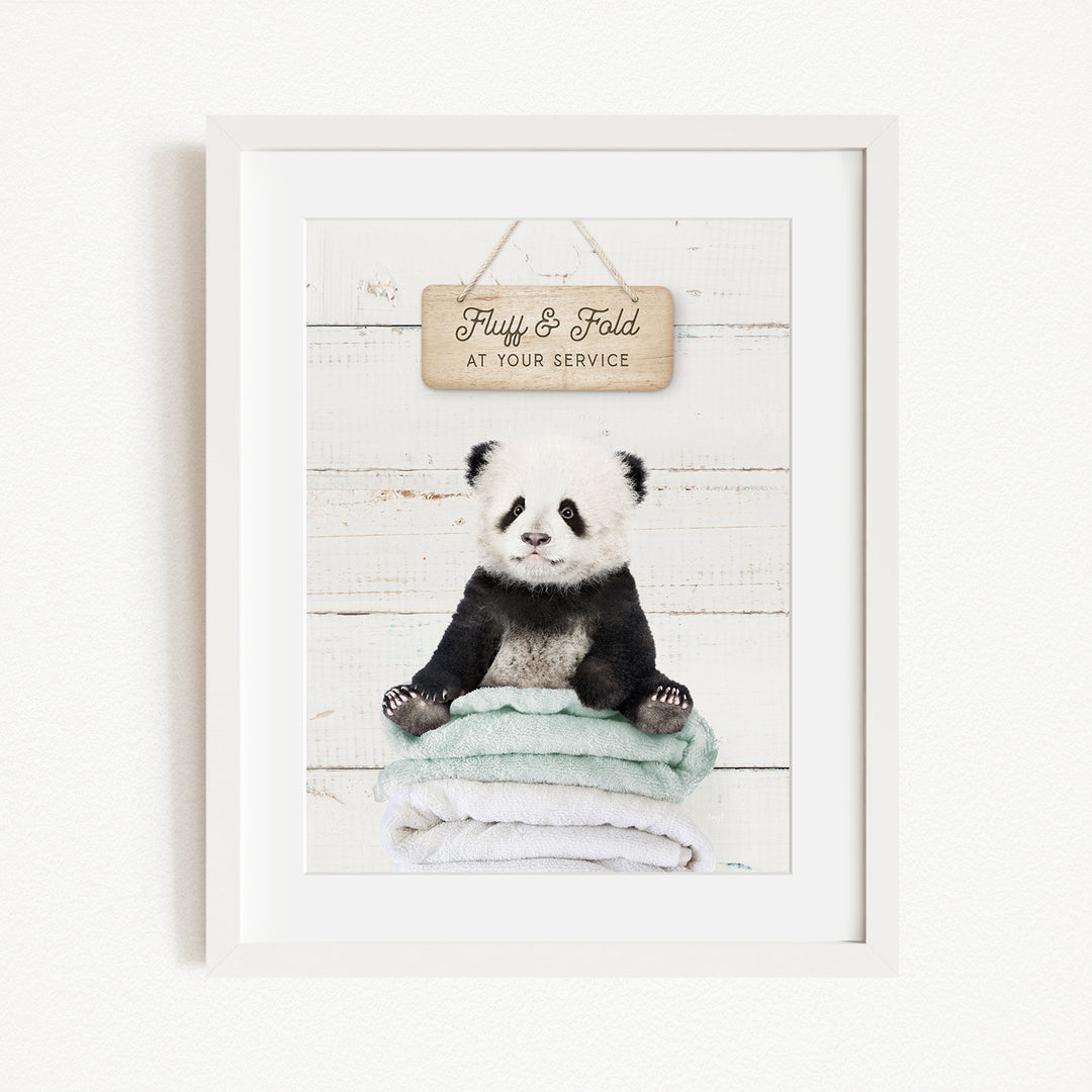 A panda bear sitting on a stack of towels, with a wooden sign above that says "Fluffy & Fold at your service".