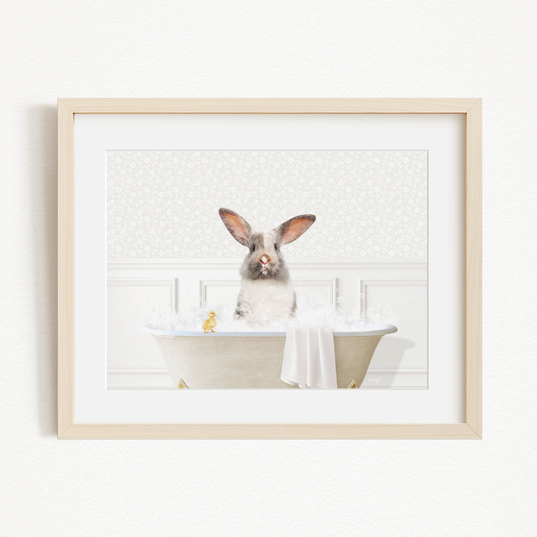 A gray and white rabbit sitting in a white bathtub filled with bubbles, with a yellow duck nearby.