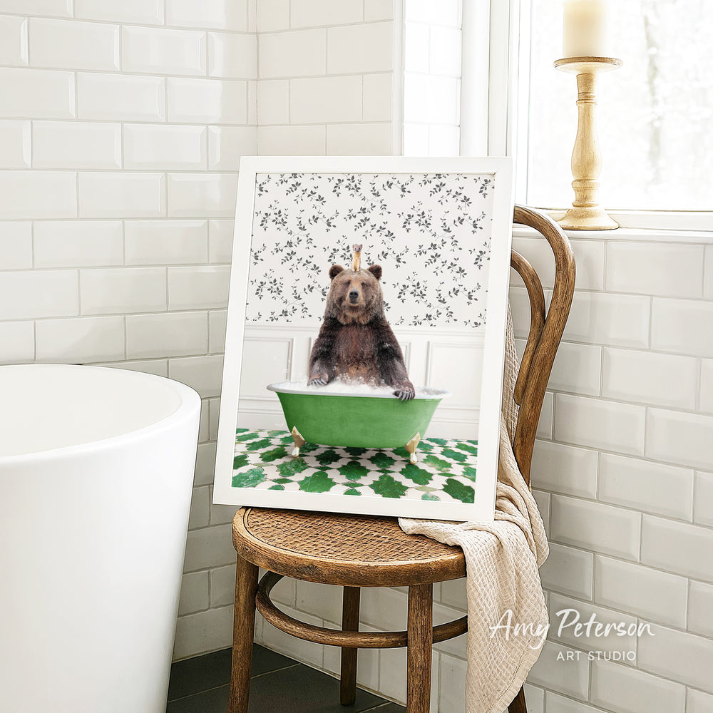 A framed image of a bear sitting in a green bathtub, surrounded by a floral pattern, is displayed on a wooden chair in a bathroom setting.