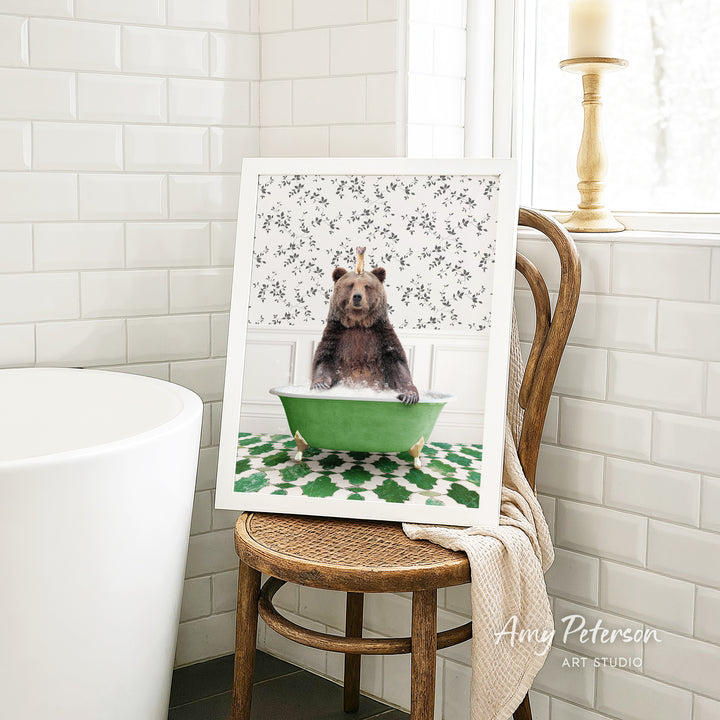 A framed image of a bear sitting in a green bathtub, surrounded by a floral pattern, is displayed on a wooden chair in a bathroom setting.