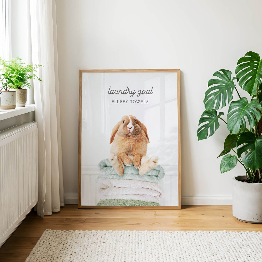 A framed poster of a rabbit sitting on a stack of fluffy towels, with the text "laundry goal" and "FLUFFY TOWELS" printed on it, is displayed on a wall in a room with a window and potted plants.