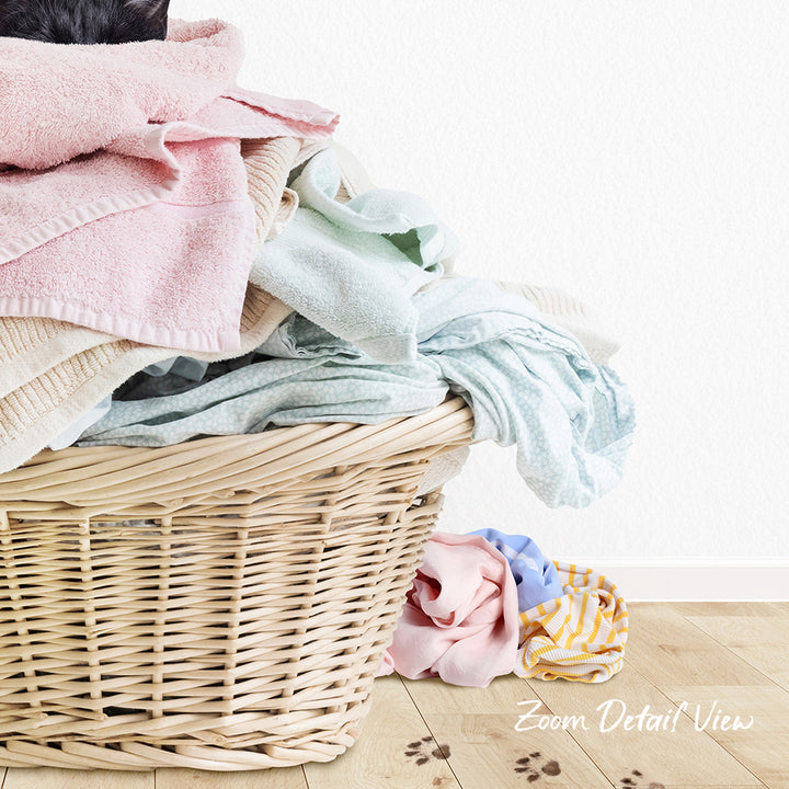 A wicker laundry basket filled with various folded clothes, including a pink towel, sits on a wooden floor.