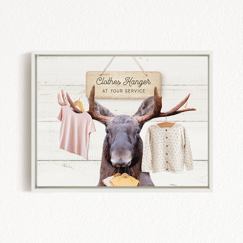 A framed image depicting a moose with antlers holding a clothes hanger, with clothing items hanging on the hanger, and a sign above the moose that reads "Clothes Hanger at Your Service".