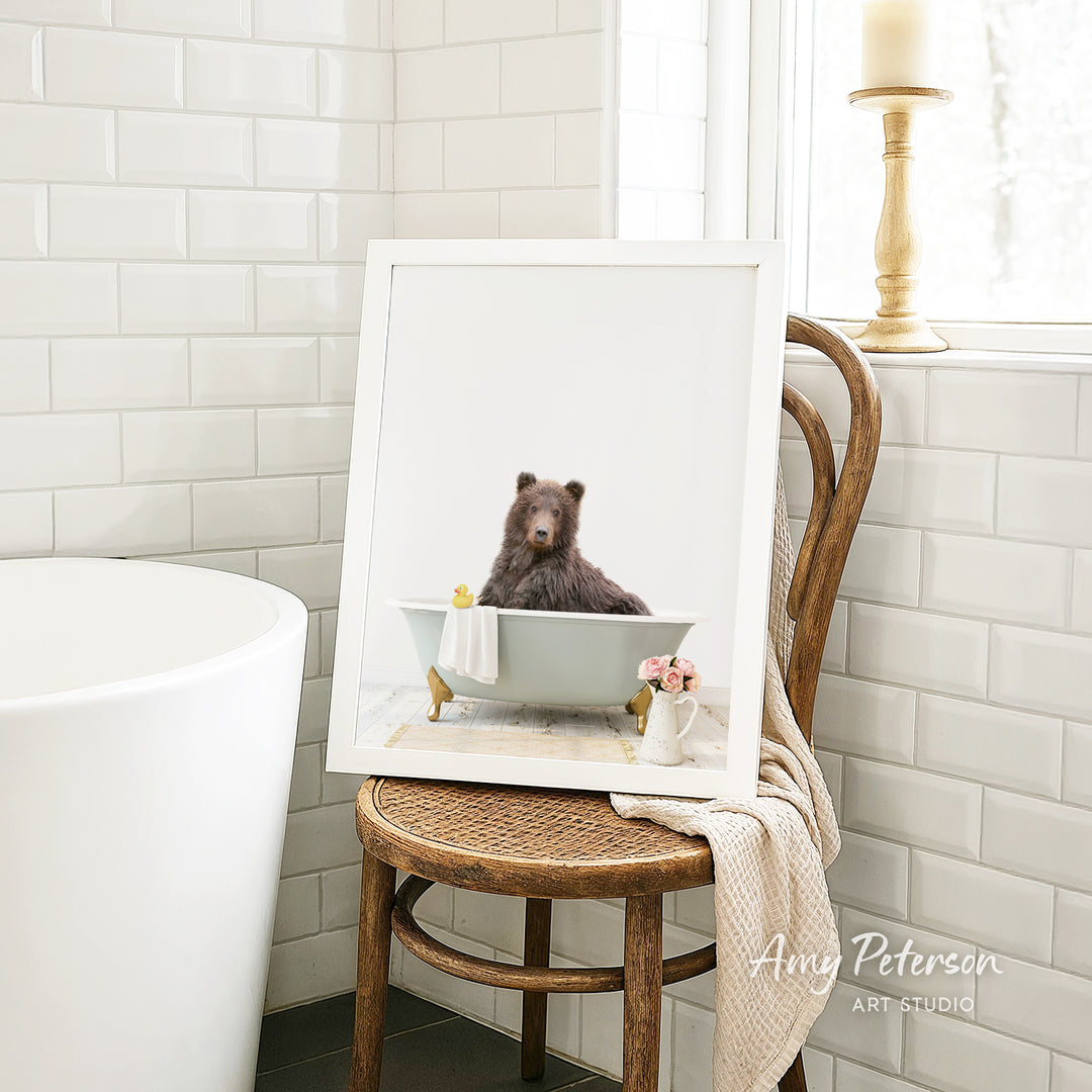 A framed image of a brown bear sitting in a bathtub, with a towel and flowers nearby, is displayed on a wooden chair in a bathroom setting.