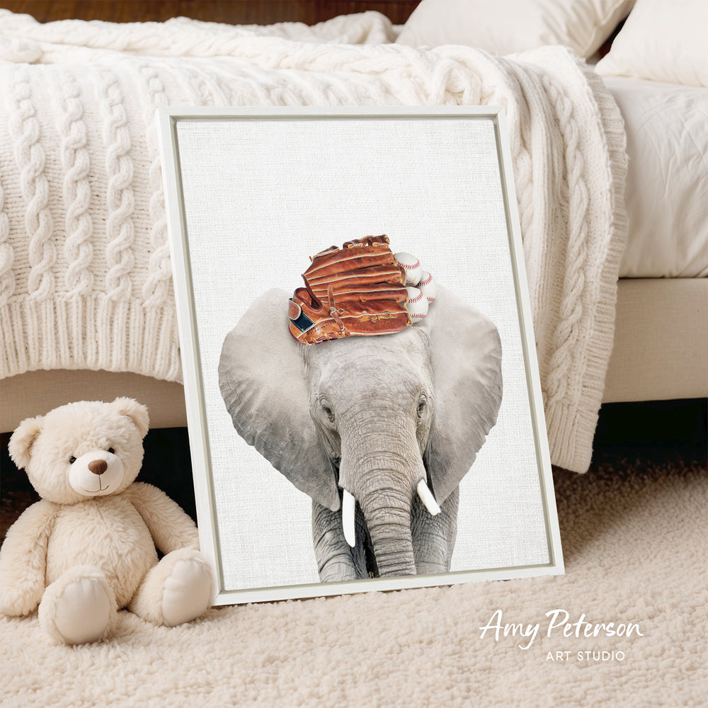 A framed print of an elephant wearing a baseball cap is displayed on a white bedspread, with a teddy bear sitting next to it.