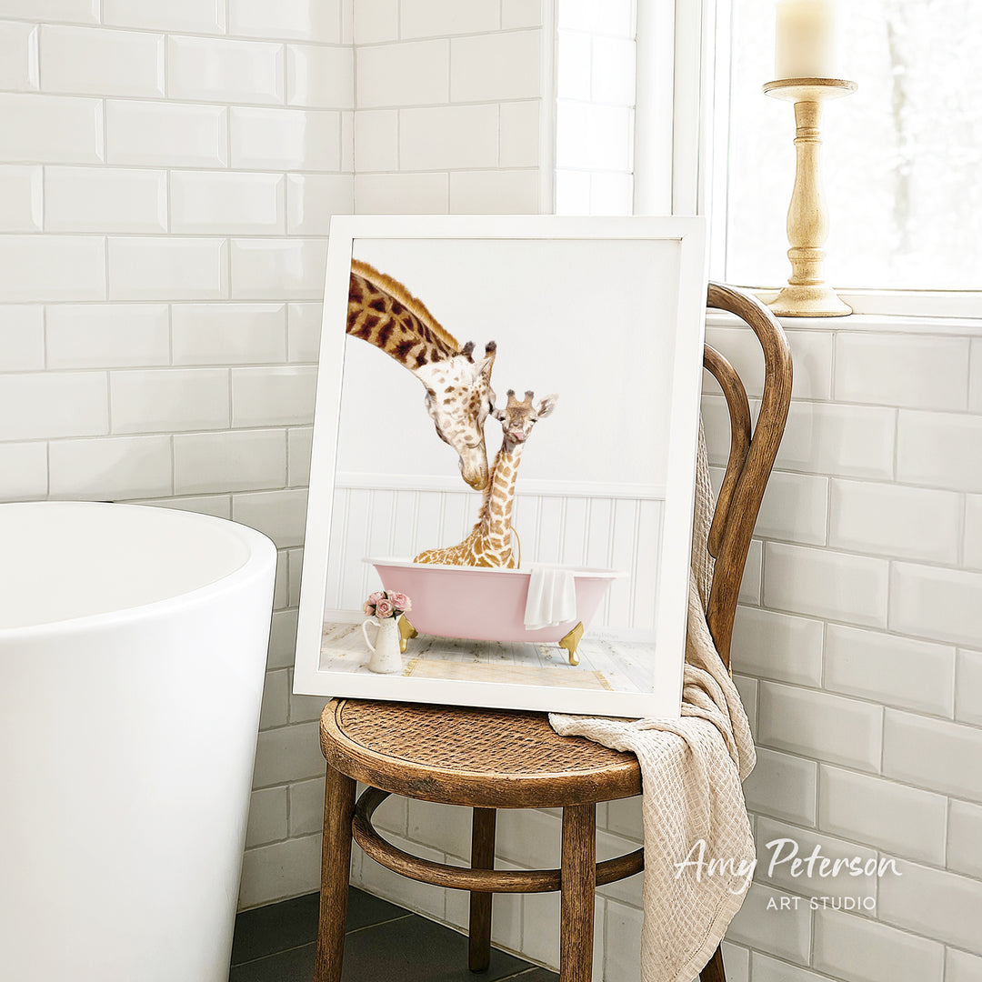 A framed image of two giraffes sitting in a pink bathtub, with one giraffe appearing to be kissing the other giraffe. The image is displayed on a wooden chair in a bathroom setting.