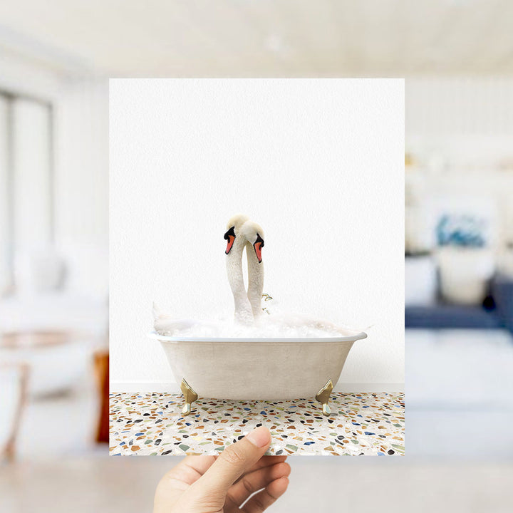 A hand holds up a photograph of two swans in a bathtub, with a blurred background suggesting a bathroom setting.