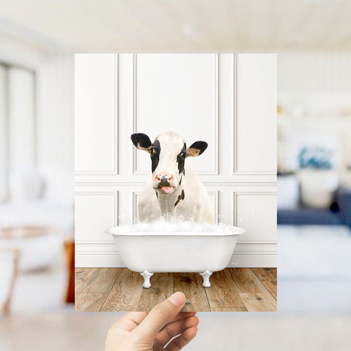A black and white cow is sitting in a white bathtub filled with water, with bubbles around it, in a white room with a wooden floor.