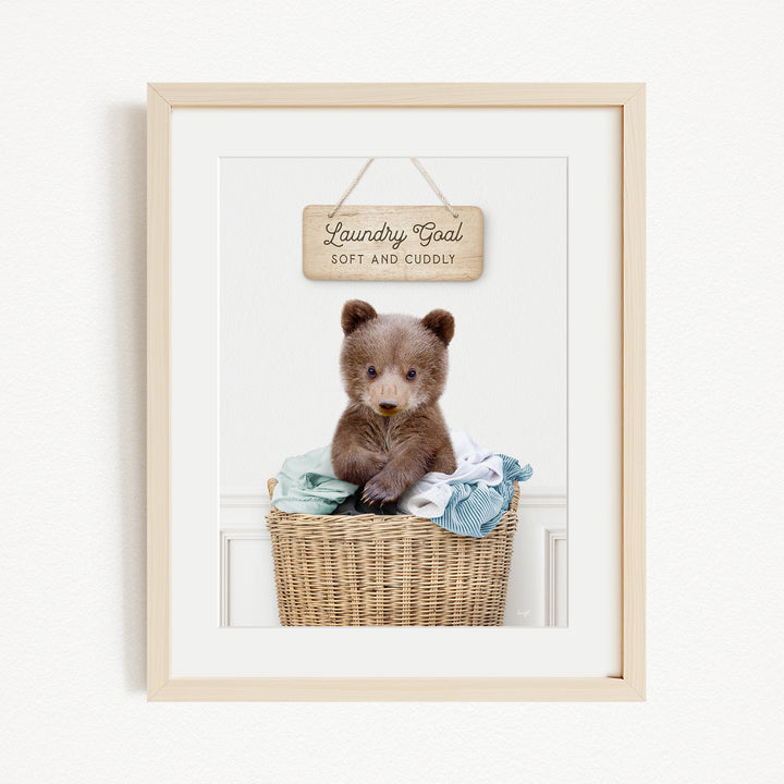 A framed photo of a baby bear sitting in a wicker laundry basket, with a wooden sign above it that says "Laundry Goal Soft and Cuddly".
