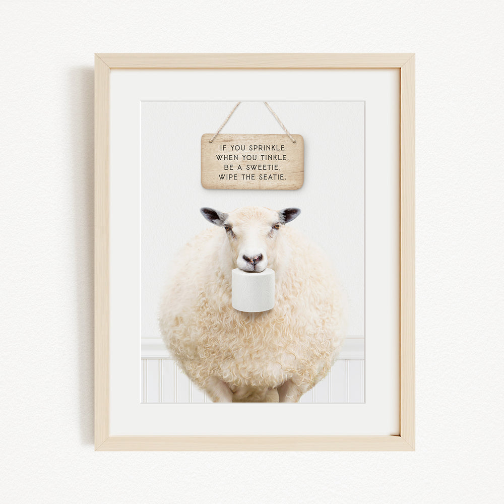 A framed image of a sheep holding a roll of toilet paper, with a wooden sign above it that reads "If you sprinkle when you tinkle, be a twinkle, wipe the seattle."