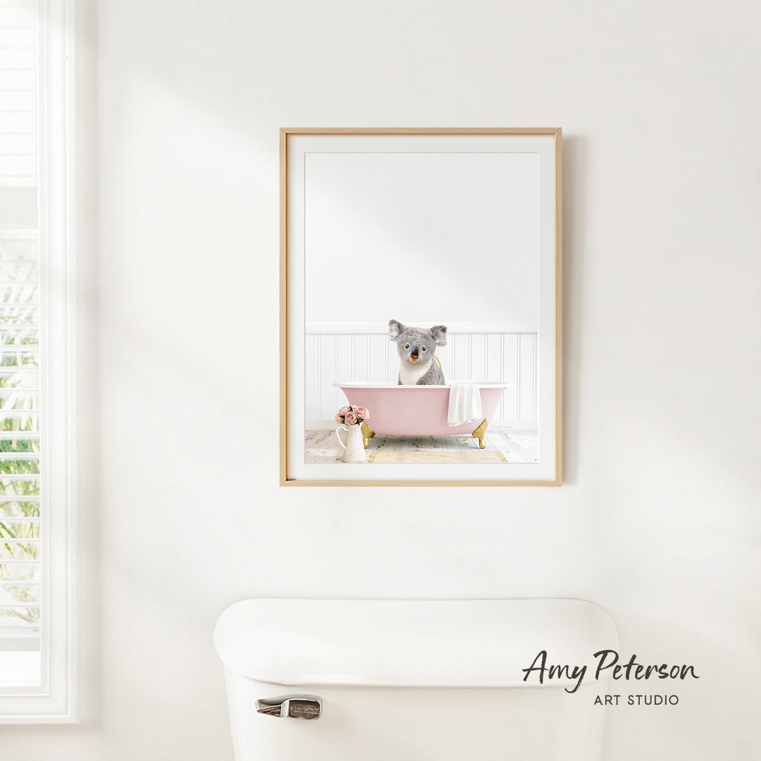 A framed image of a koala sitting in a pink bathtub, with a vase of flowers and a white toilet in the foreground.