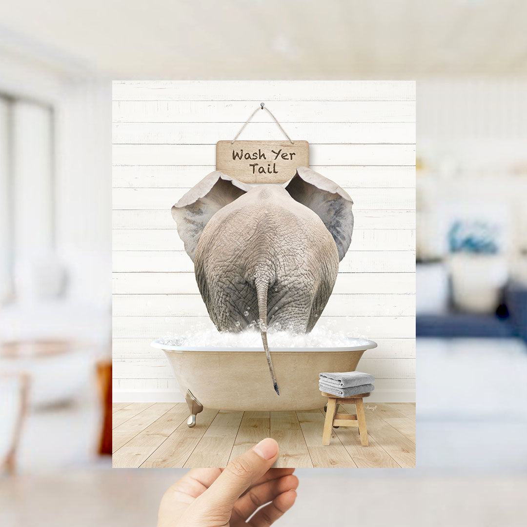 A hand holds up a framed image depicting an elephant sitting in a bathtub, with a sign above it that says "Wash Your Tail".