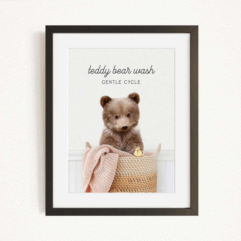 A framed image of a teddy bear sitting in a woven basket, with a blanket and a small toy duck inside.