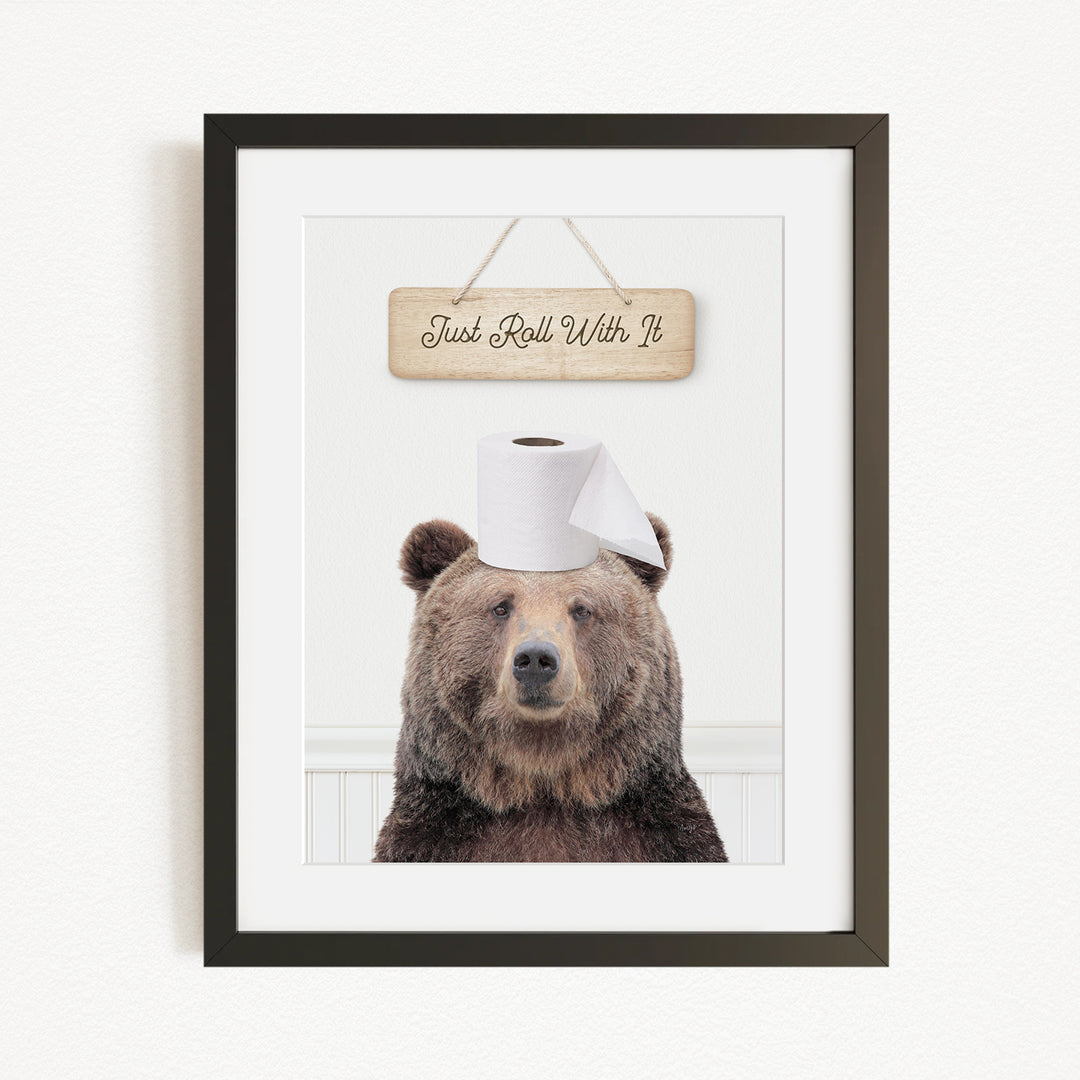 A framed image of a brown bear wearing a toilet paper hat, with a wooden sign above it that says "Just Roll With It".