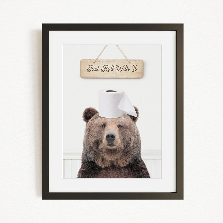 A framed image of a brown bear wearing a toilet paper hat, with a wooden sign above it that says "Just Roll With It".