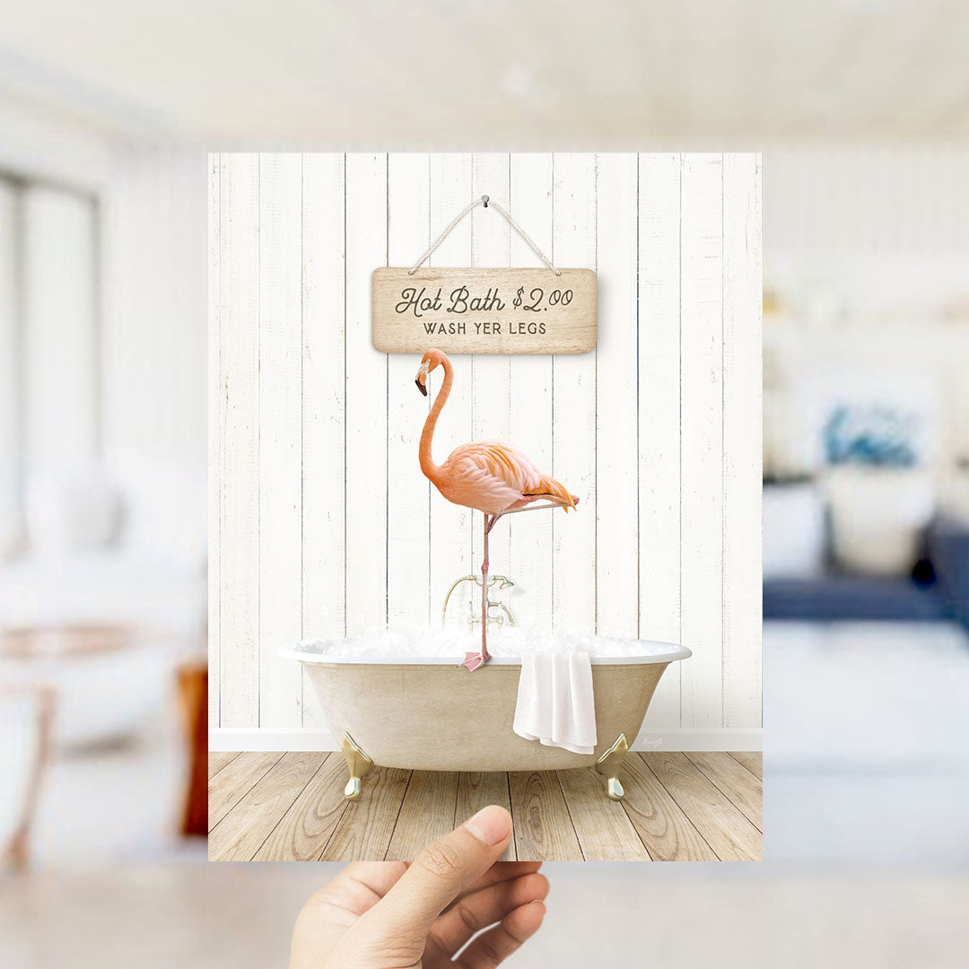 A hand holding a framed image of a pink flamingo standing in a bathtub, with a sign above it that says "Hot Bath $2.00 WASH YER LEGS".