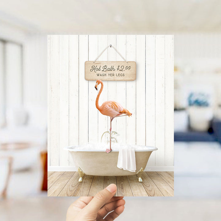 A hand holding a framed image of a pink flamingo standing in a bathtub, with a sign above it that says "Hot Bath $2.00 WASH YER LEGS".