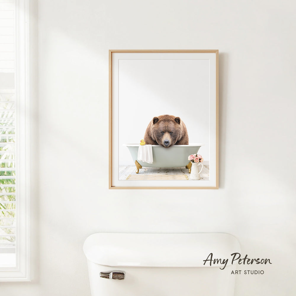 A framed image of a brown bear sitting in a bathtub, with a vase of flowers and a white toilet visible in the background.