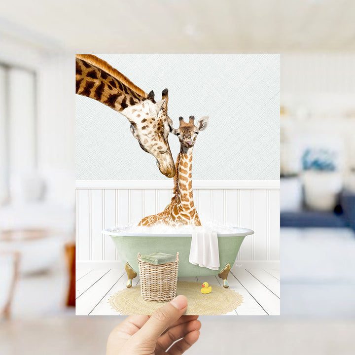 A hand holds up a framed image depicting two giraffes in a bathtub, with one giraffe appearing to be a baby giraffe.