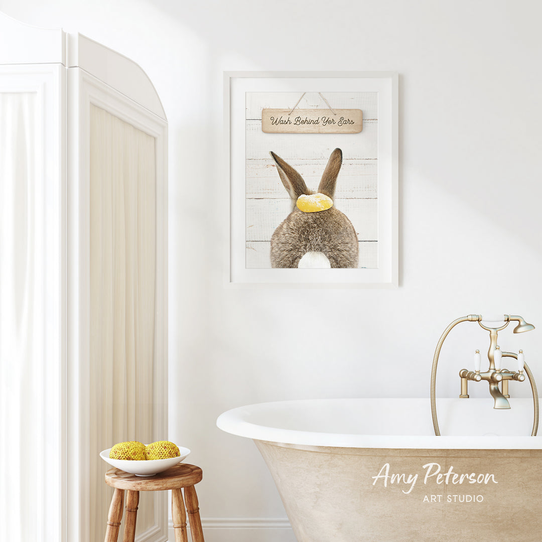 a bathroom with a framed picture of a rabbit on the wall, a wooden stool, a bowl of fruit, and a bathtub with a faucet.