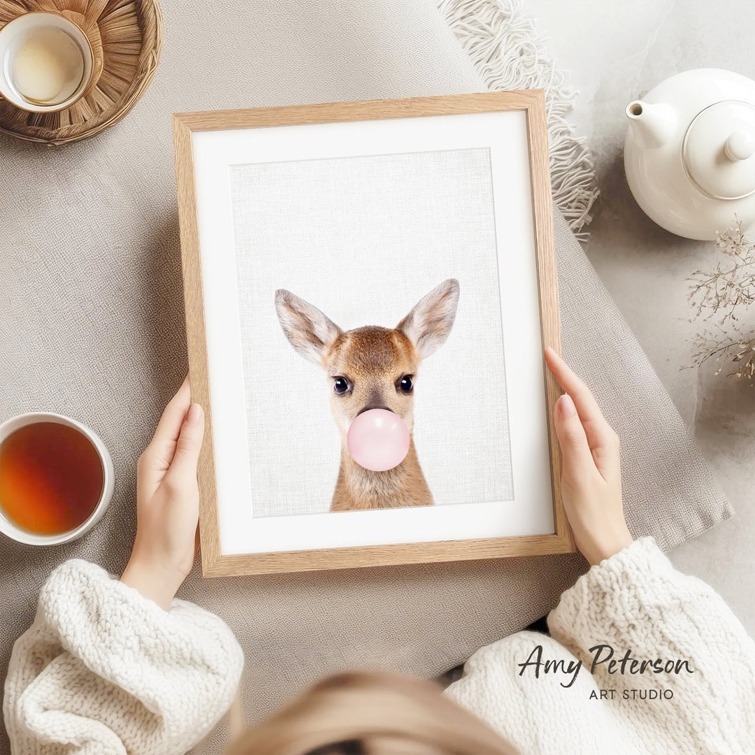 A person is holding a framed picture of a deer with a bubble gum bubble in its mouth, surrounded by various objects such as a teapot, a cup, and a bowl.