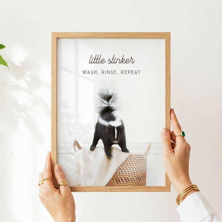 A person's hands holding a framed picture of a skunk, with the text "little stinker" and "WASH, RINSE, REPEAT" printed on the frame.