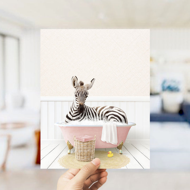 A zebra is sitting in a pink bathtub filled with water, surrounded by a yellow rubber duck and a white towel.